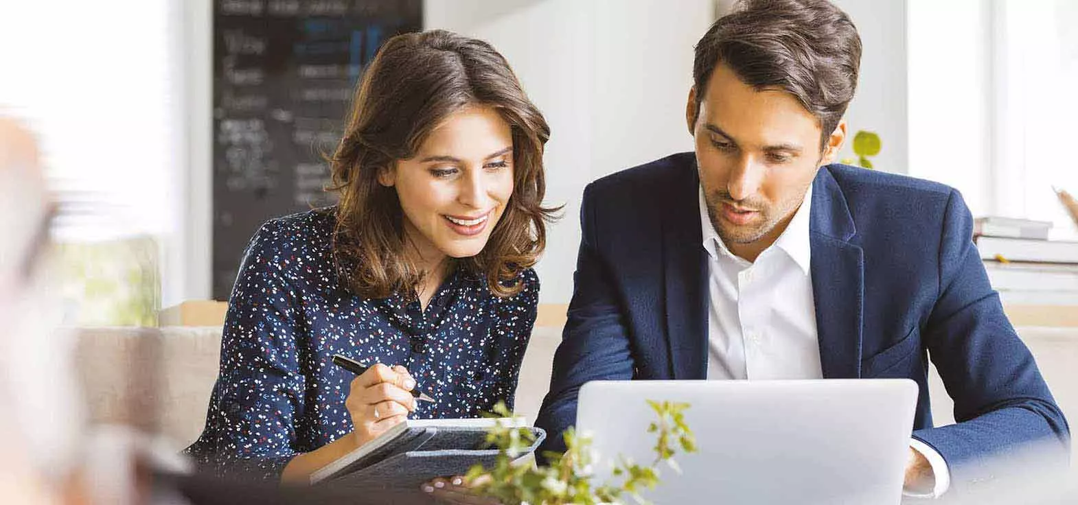 An executive woman is taking notes as an executive man is showing her results on his laptop